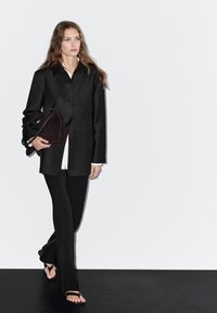 Black button-up jacket over a white blouse, paired with black trousers and flat sandals. Model carries a dark brown handbag.