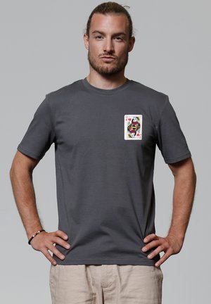 Mann mit zurückgebundenem Haar, der ein graues T-Shirt mit einer Spielkarte der Herzkönigin auf der Brust trägt und mit den Händen in den Hüften dasteht.