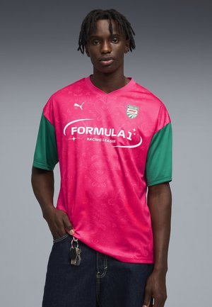 Jonge man met dreadlocks die een roze en groene Formula 1 Racing League-trui draagt, sleutels vasthoudt en staat tegen een effen grijze achtergrond.