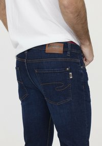 Jean en denim bleu foncé avec un patch en cuir à la taille, couture orange contrastante, et deux poches arrière avec des coutures décoratives.