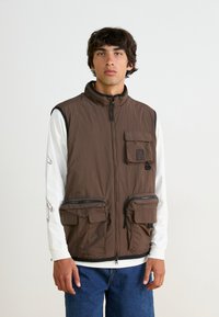 Brown sleeveless vest made of nylon, features a high collar, front zip closure, one chest pocket, and two large zippered pockets.