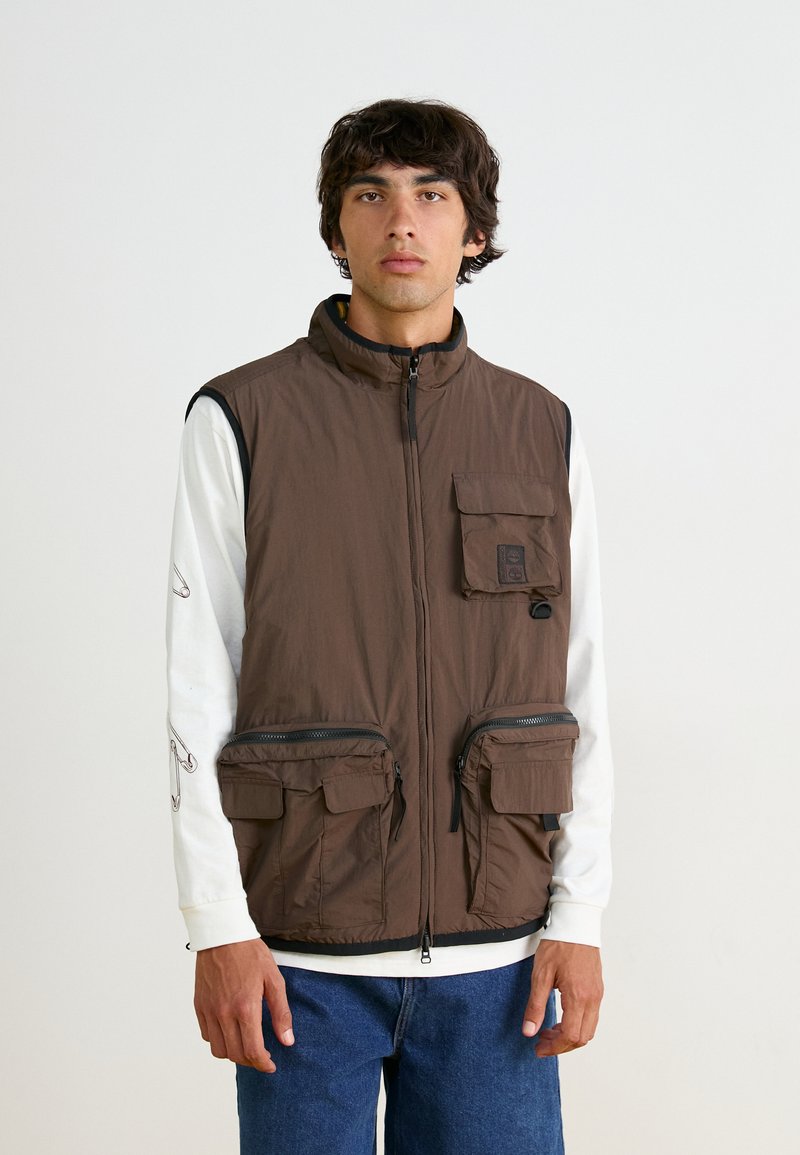 Brown sleeveless vest made of nylon, features a high collar, front zip closure, one chest pocket, and two large zippered pockets.