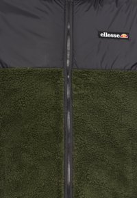Chaqueta de forro polar verde oliva con una sección superior negra, cierre de cremallera y logotipo bordado de "ellesse" en el pecho. Textura suave y diseño cálido.