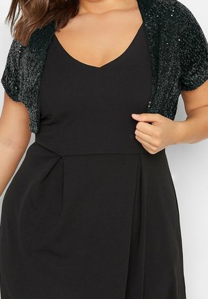 Femme portant une robe noire sans manches avec un décolleté en V et un boléro noir à sequins à manches courtes, tenant le boléro d'une main.
