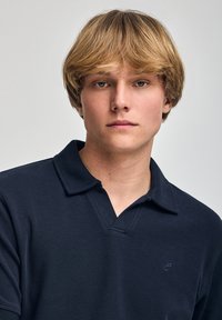 Camisa de polo azul marino con una tela texturada, cuello corto y un logo sutil en el lado izquierdo. El modelo muestra una expresión neutral.