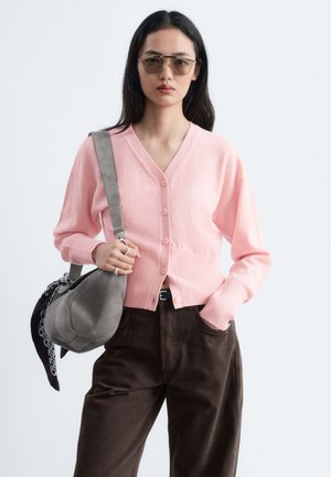 Femme portant un cardigan rose boutonné, un pantalon foncé, des lunettes de soleil et un sac à bandoulière gris avec un foulard à motif attaché.