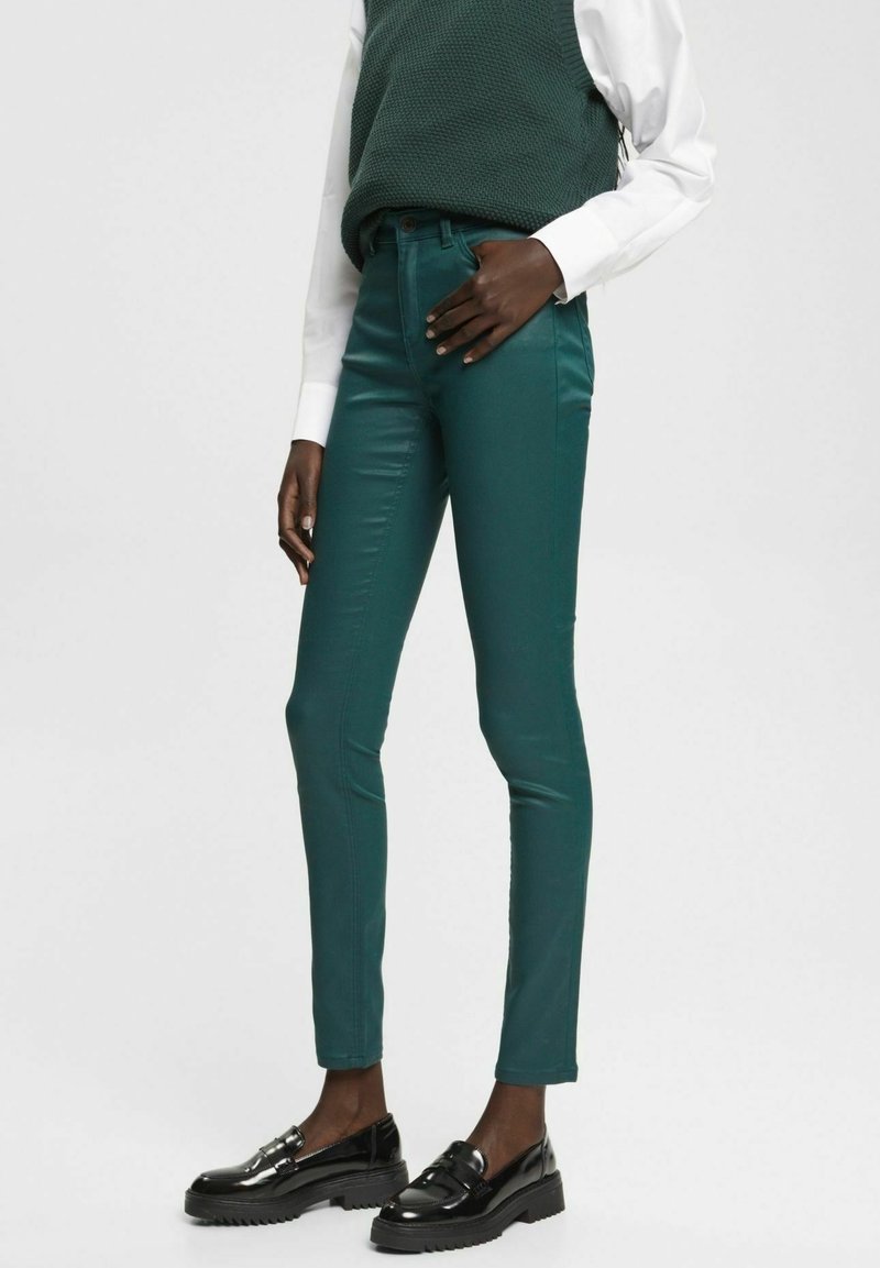 Pantalons ajustés teal à finition brillante, mettant en valeur une taille mi-haute et des poches latérales, associés à des mocassins noirs brillants.