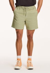 Světle zelené kraťasy s elastickým páskem a šňůrkou. Obsahují boční kapsy a mají ležérní design, doplněné oranžovými vyššími teniskami.