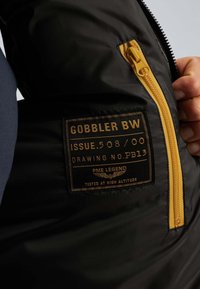 Unutrašnjost crne jakne sa žutim patentnim zatvaračem i etiketom na kojoj piše "Gobbler BW, PME Legend, testirano na visokoj nadmorskoj visini."