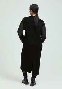 Schwarzes Kleid mit langen, durchsichtigen Ärmeln, gerader Silhouette, Schlitz im Rücken und rundem Ausschnitt. Getragen mit schwarzen Kampfschuhen.