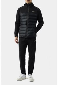 Zwarte gewatteerde jas met een ritssluiting aan de voorkant, een contrasterend glad bovenstuk, gecombineerd met zwarte broek en witte sneakers. Getextureerde accenten op de jas.