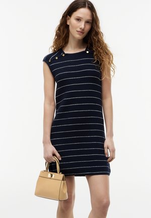 Jeune femme aux cheveux ondulés portant une robe sans manches rayée bleu marine, tenant un sac à main beige avec des anses supérieures.