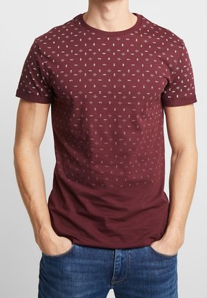 Mann trägt ein kurzärmeliges bordeauxrotes T-Shirt mit kleinen weißen geometrischen Mustern und blaue Jeans, Hände in den Taschen, steht vor einem einfarbigen Hintergrund.