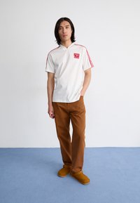 Polo branca de mangas curtas com riscas vermelhas e logotipo, combinada com calças castanhas e tamancos castanhos. Modelo está de pé sobre um tapete azul claro.
