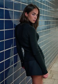Young woman in black turtleneck and skirt leaning against blue tiled wall, looking over her shoulder in a corridor.