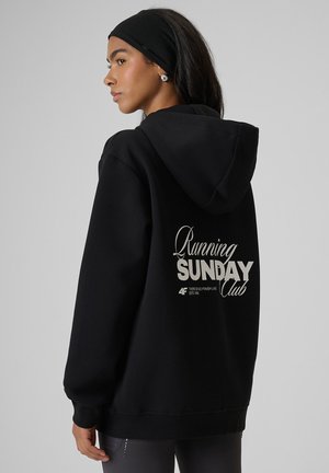 Kobieta w czarnej bluzie z kapturem z napisem "Running Sunday Club" na plecach, z czarną opaską na głowie i srebrnymi kolczykami, patrzy w lewo.