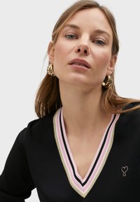 Crna v-neck pulover s višestrukom prugastom ovratnikom u ružičastoj, zlatnoj i crnoj boji; ukrašen malim detaljem u obliku srca od kristala.