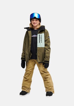 Junge Person trägt eine grüne und beige Schneejacke, beige Schneehosen, schwarze Handschuhe und blaue Skibrille auf einer schwarzen Mütze.