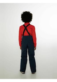 Maglia rossa a maniche lunghe con colletto alto indossata sotto bretelle nere, abbinata a pantaloni da sci blu scuro con gamba dritta e tasche posteriori.