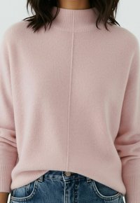 Pull à col montant côtelé rose pâle, porté rentré dans un jean en denim bleu, sur une personne aux cheveux foncés mi-longs.