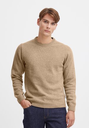 Pull en tricot beige avec un col rond, des bords-côtes aux poignets et à l'ourlet. Texture lisse, coupe ajustée. Porté sur un pantalon en denim foncé.