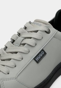 Ανοιχτό γκρι αθλητικό παπούτσι με υφασμάτινα κορδόνια, διακριτικό λογότυπο, λάστιχο σόλα και λείο δερμάτινο επάνω μέρος. Διαθέτει αντίθετες μαύρες πινελιές και ραφές.