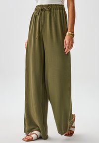 Pantaloni verdi a gamba larga in tessuto lavorato, con vita elastica e coulisse. Abbinati a sandali aperti.