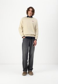 Beige crew neck pulóver, egy kis piros logóval, sötétkék ing fölött. Szürke farmer és barna cipő egészíti ki az öltözetet.