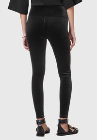 Persona che indossa leggings di velluto nero e sandali neri con grandi fibbie dorate, mostrata di spalle su uno sfondo semplice.