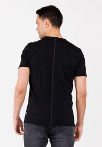 Hombre vestido con una camiseta negra de manga corta con una costura central visible en la parte posterior y unos jeans gris oscuro, de espaldas contra un fondo blanco liso.