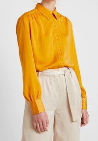 Žlutá košile z lesklého hedvábí s dlouhými rukávy, knoflíkovou légou, nafouklými rukávy a knoflíkovými detaily; doplněná světlými béžovými kalhotami širokých nohavic s páskem.