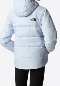 Jasnoniebieska kurtka puchowa z kapturem i czarnym logo "The North Face" na górnej części pleców, noszona z czarnymi spodniami na jednolitym tle.