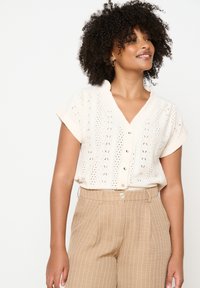 Balta trumpa rankovėmis bluzonė su sudėtingais aklumo raštais, V formos iškirpte ir mygtukų uždarymu, derinama su smėlio spalvos juostuotais kelnių.