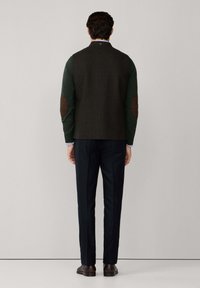 Blazer in misto lana di colore verde scuro con retro testurizzato e toppe marroni sui gomiti, abbinato a pantaloni blu navy e scarpe marroni, visto da dietro.