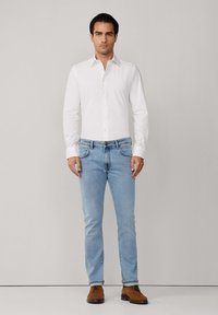 Muškarac stoji uz jednobojnu pozadinu, odjeven u bijelu košulju s dugim rukavima, svijetloplave traperice i smeše cipele na vezivanje.