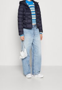 Marinblå puffjacka med huva, randig blå och beige tröja, vida ljusblå jeans, vita sneakers och en liten vit handväska.