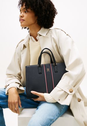 Γυναίκα με σγουρά μαλλιά που φοράει μπεζ παλτό τρεντσκότ και πλεκτό πουλόβερ, κρατώντας μαύρη τσάντα χειρός με κόκκινες και λευκές ρίγες ενώ είναι καθιστή.