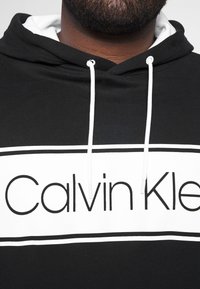 Felpa nera in misto cotone, con una scatola logo bianca recante la scritta "Calvin Klein", cordini bianchi e una fodera del cappuccio bianca a strati.