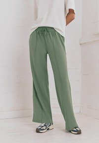 Pantaloni della tuta leggeri e verdi con un elastico in vita, vestibilità rilassata e cuciture laterali. Abbinati a sneakers a fantasia e una maglietta bianca.