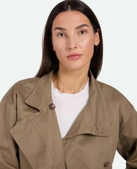 Gabardina beige con cuello de muesca, diseño de doble botonadura y grandes botones marrones. Textura suave, llevada sobre una camisa blanca.