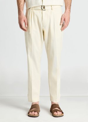 Homme portant un pantalon plissé couleur crème et des sandales marron à bout ouvert, debout devant un fond blanc uni.
