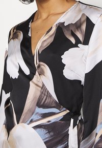 Top enveloppant noir avec de grands motifs floraux en crème et marron, décolleté en V et détail de lien sur le côté. Matière en soie lisse.