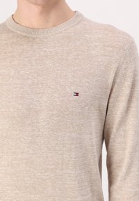 Beige neulepaita, jossa on pyöreä kaula-aukko ja vasemmalla rintapielellä pieni punaisella, valkoisella ja sinisellä brodeerattu logo. Pehmeä tekstuuri.