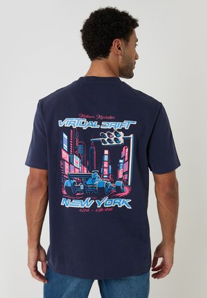 Mand iført en navyblå t-shirt med neonstil grafik af racerbiler og bylandskab mærket "Virtual Drift New York 42nd-49th Street."