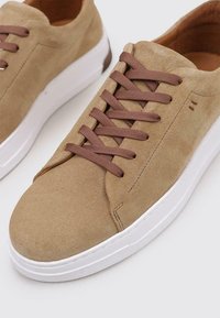 Zapatillas de ante beige con una textura suave, que presentan cordones marrones y una suela de goma blanca. Detalle de costura lateral sutil.