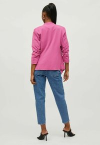 Rosa blazer med lange ermer og en lett utskjært kant, kombinert med blå denimjeans og svarte hæl-sandaler. Glatt tekstur.