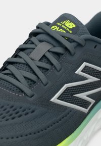 Chaussure de sport gris foncé avec dessus en mesh, lacets plats gris, accents réfléchissants et logo vert vif. Caractérisée par une semelle texturée.