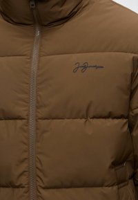 Veste matelassée marron avec fermeture éclair à l'avant et un petit logo sombre sur la partie gauche de la poitrine.