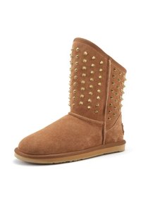 Botín de ante beige con tachuelas de metal dorado en un lado, suela plana de goma, punta redondeada y textura suave. Presenta costuras reforzadas.