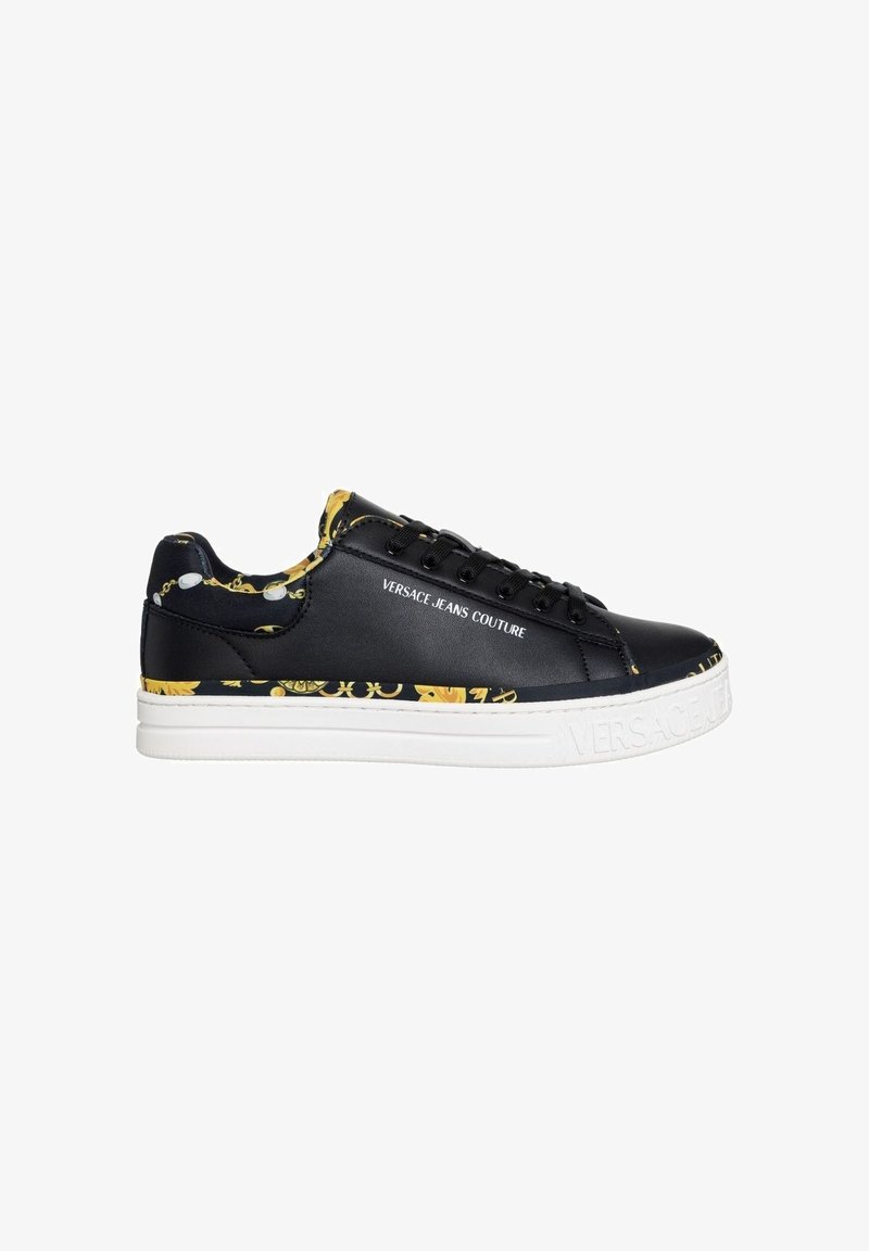 Baskets en cuir noir avec des accents en motif chaîne dorée, semelle blanche, design à lacets et logo sur le côté. Texture lisse dans l'ensemble.